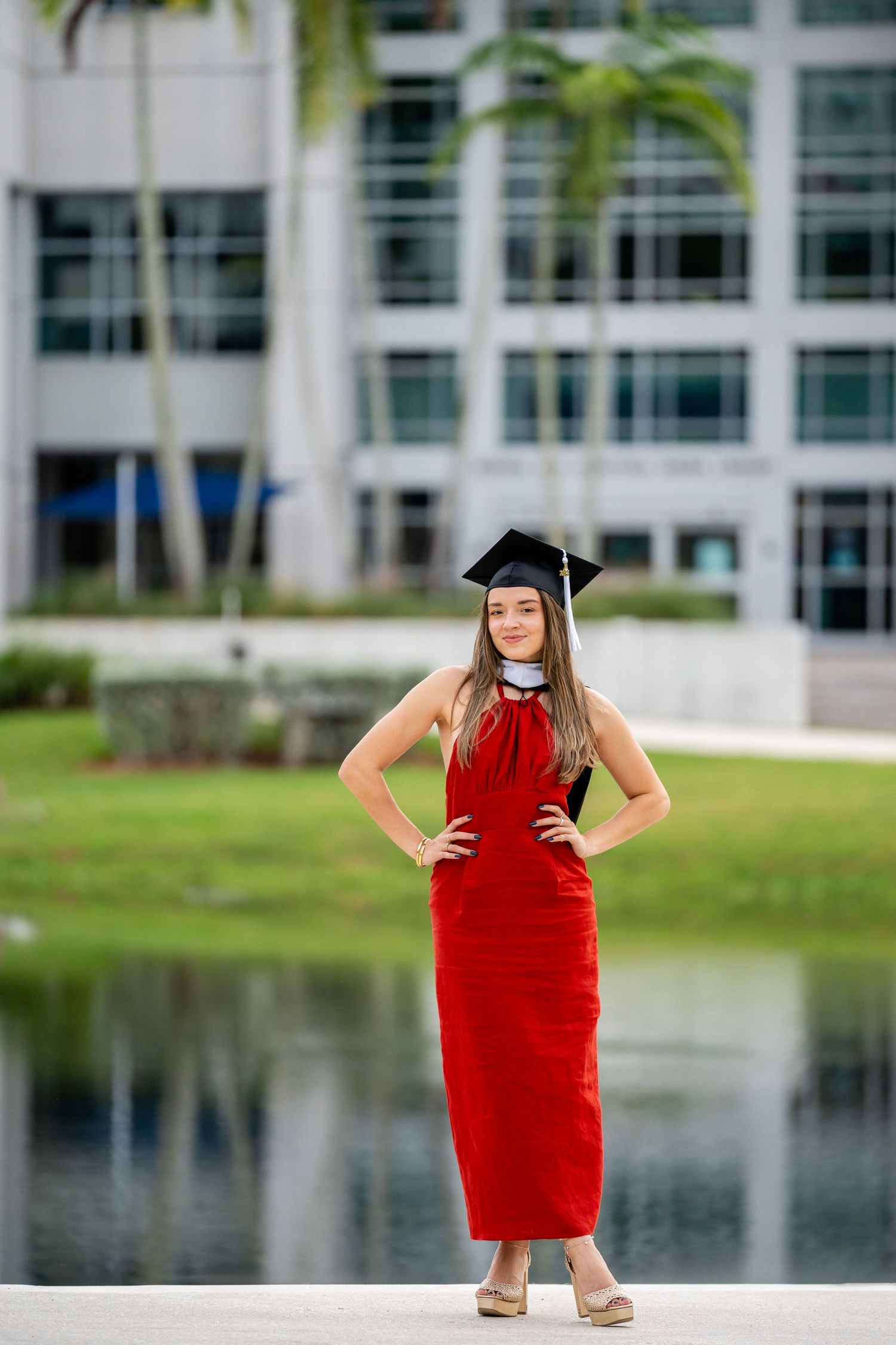 Graduate in red dress and cap poses by pond with university building in background.