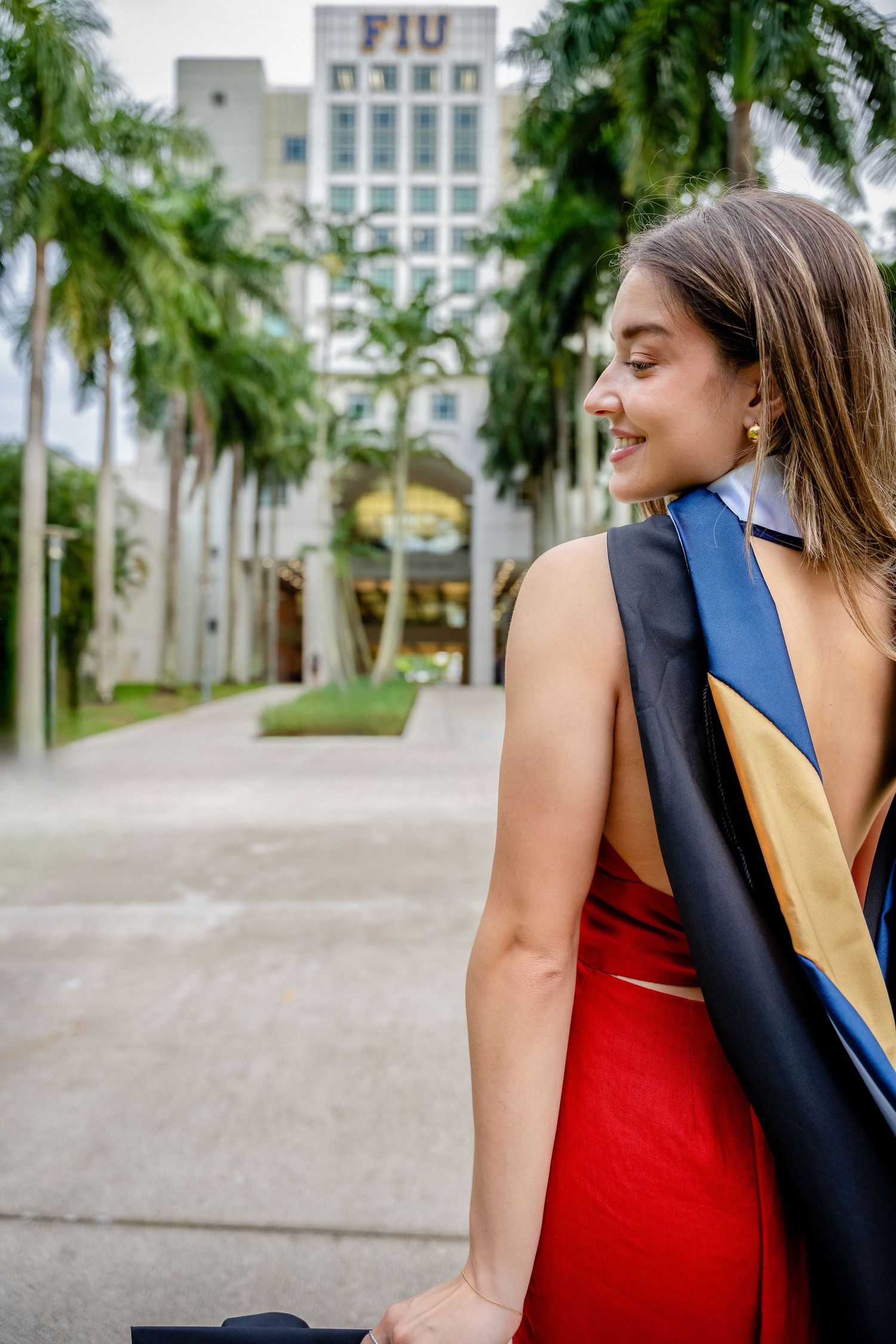 Graduate wearing red dress and academic stole poses in palm tree-lined path.