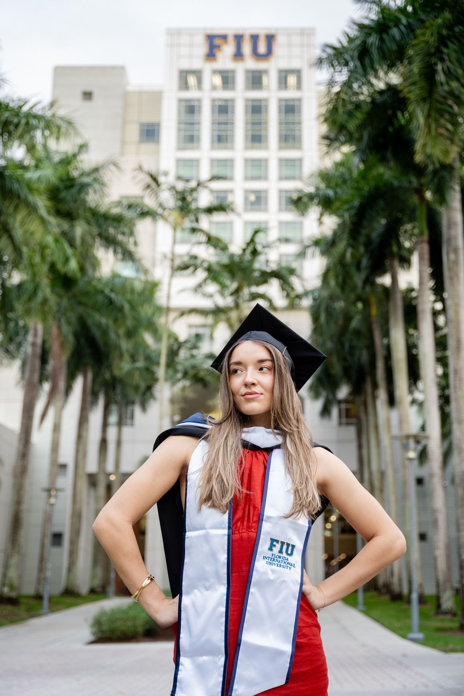 Graduate in red dress and academic regalia poses in front of FIU building with palm trees.