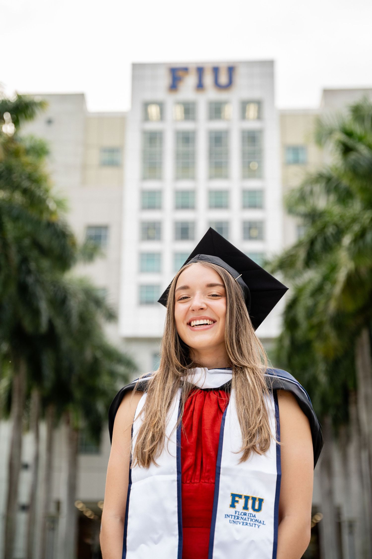Graduate in cap and gown smiles joyfully in front of FIU building with palm trees.