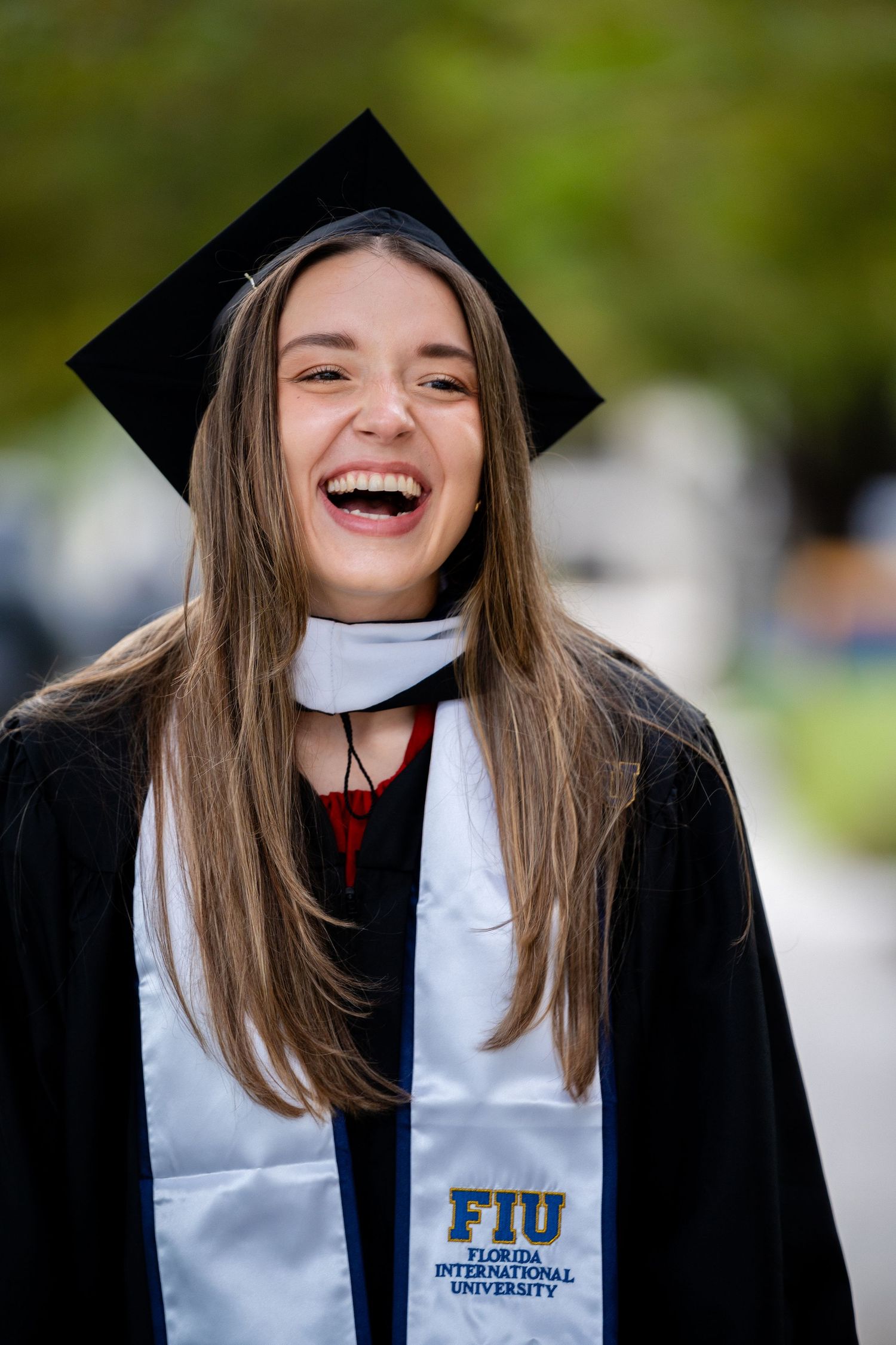 Graduate in cap and gown laughs joyfully during outdoor graduation photoshoot.