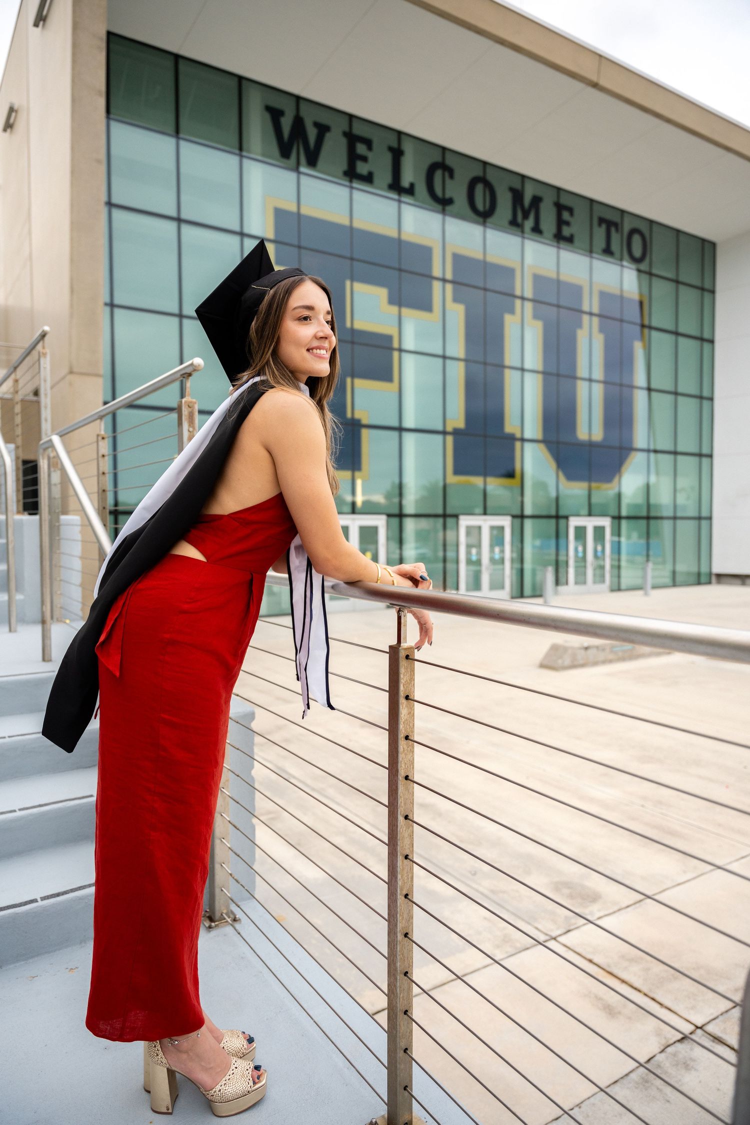 Graduate in red dress and cap poses by Welcome to FIU sign.
