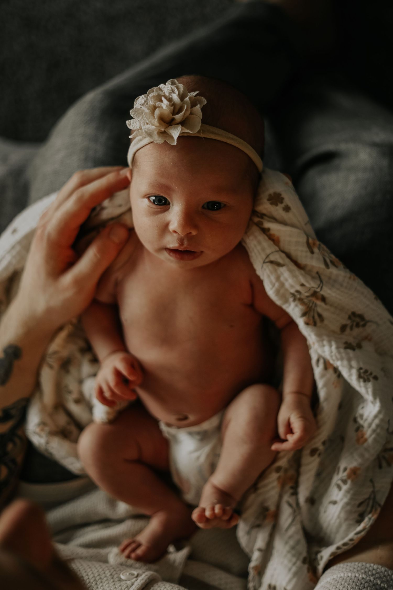 Baby wearing a floral headband lies on textured blankets in moody lighting.