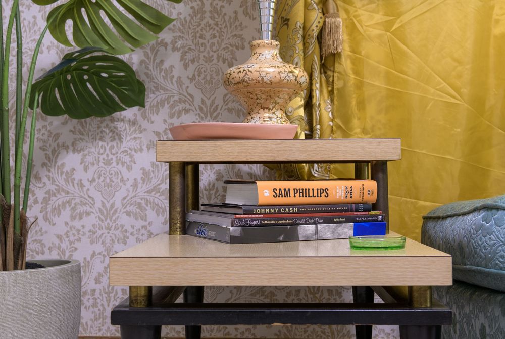 A wooden side table displays stacked books between decorative monstera plants and yellow curtains in a modern living room.