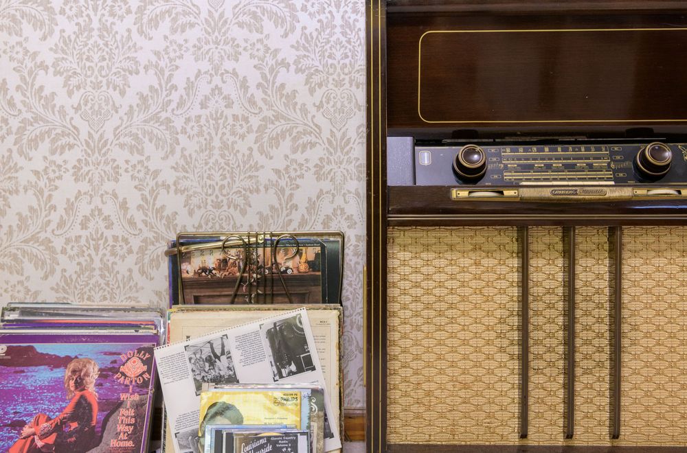 Vintage radio receiver with gold mesh speaker grill displayed next to vinyl records against floral wallpaper.
