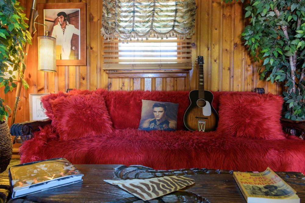 A cozy rustic living room with wood paneling, red plush couch, acoustic guitar and decorative window treatment.