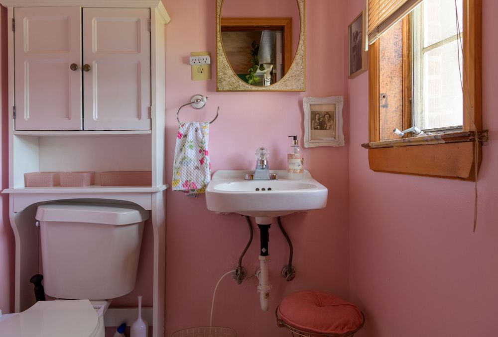 A vintage pink bathroom interior with pedestal sink, white cabinet, and small window letting in natural light.