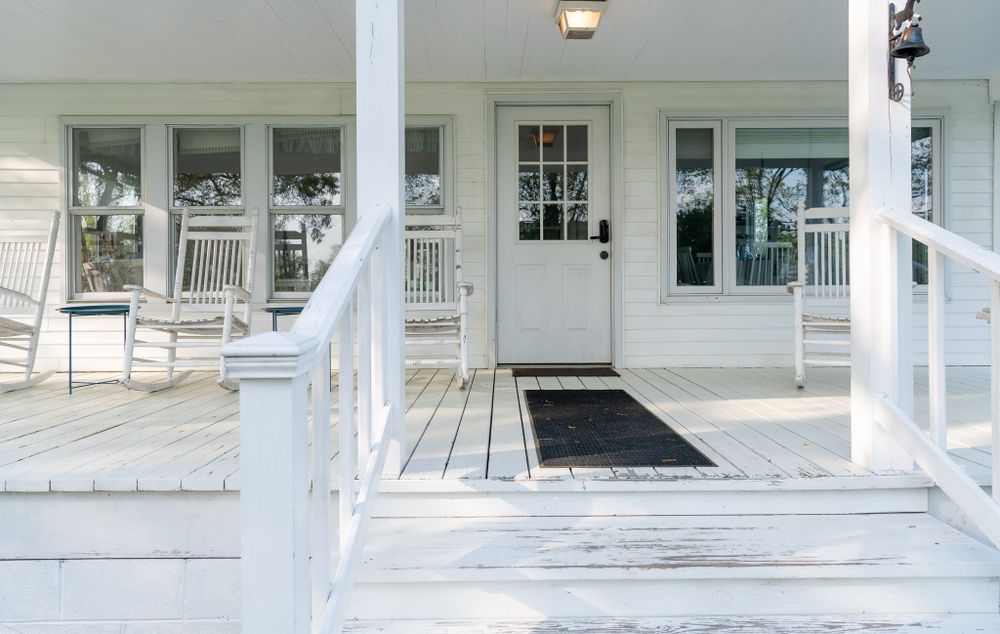 White wooden porch with rocking chairs and a black doormat leading to a classic front door entrance.