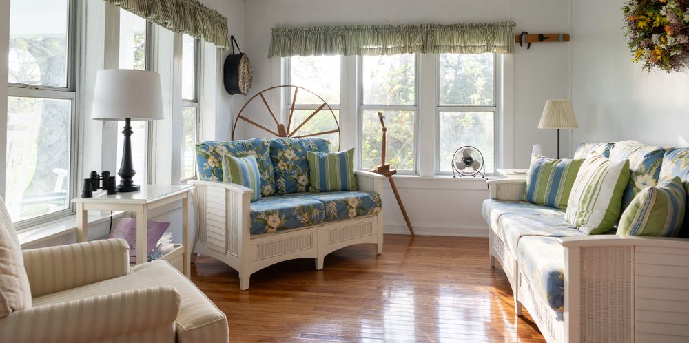 Bright sunroom with white wicker furniture, blue and green throw pillows, and hardwood floors in a coastal style setting.