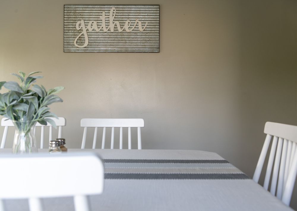 A minimalist dining area with white chairs, striped table runner, and 'gather' wall sign above green potted plant.
