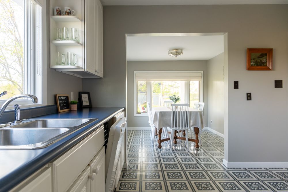 Modern kitchen with white cabinets and dark countertops opens to bright dining area with patterned tile flooring.