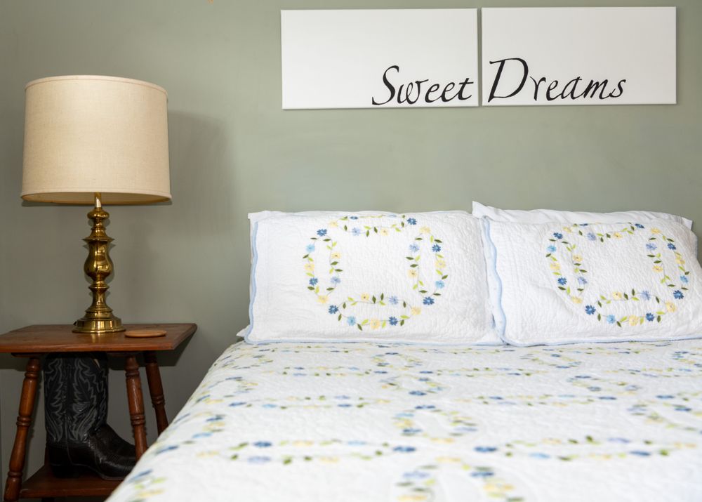 A cozy bedroom with white bedding featuring floral patterns, a wooden side table, and 'Sweet Dreams' wall art.