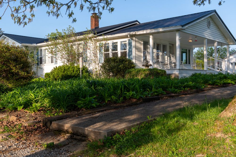 White ranch style home with wrap-around porch sits atop grassy hill with stone path and garden landscaping.