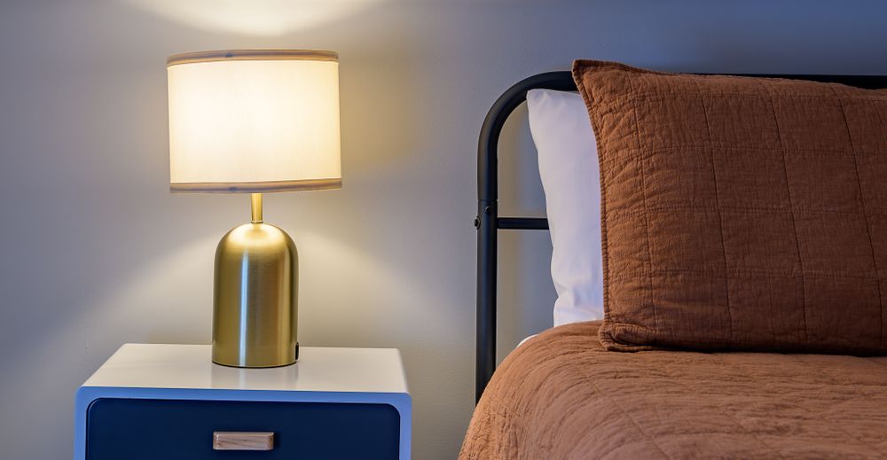 Modern gold table lamp with white shade sits on blue nightstand beside tan bed comforter and brown pillow.