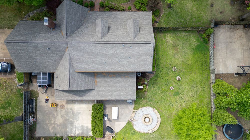 Aerial view of a residential house with gray roof and spacious backyard featuring circular patio element.