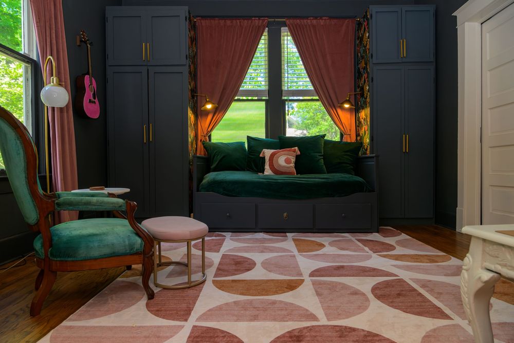 A cozy reading nook features a deep green velvet sofa against dark built-ins with a pink geometric area rug.