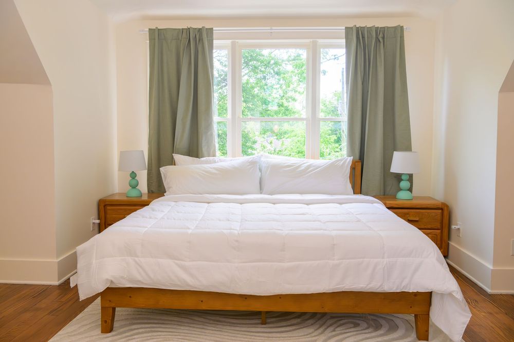 Bright bedroom with white bedding, wooden furniture, and bay window with sage green curtains overlooking garden view.