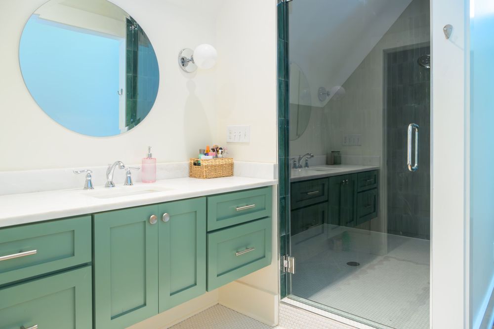 Modern bathroom with sage green vanity, round mirror, and glass walk-in shower under sloped ceiling.