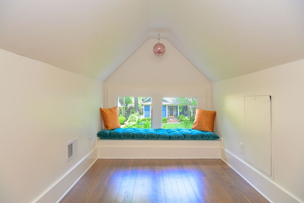 Cozy attic reading nook with built-in bench seating, orange pillows and window overlooking greenery.