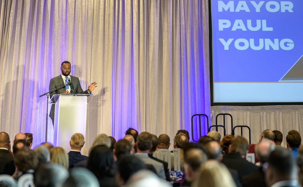 A speaker addresses a large audience from a podium at an event with 'Mayor Paul Young' displayed on a projection screen.