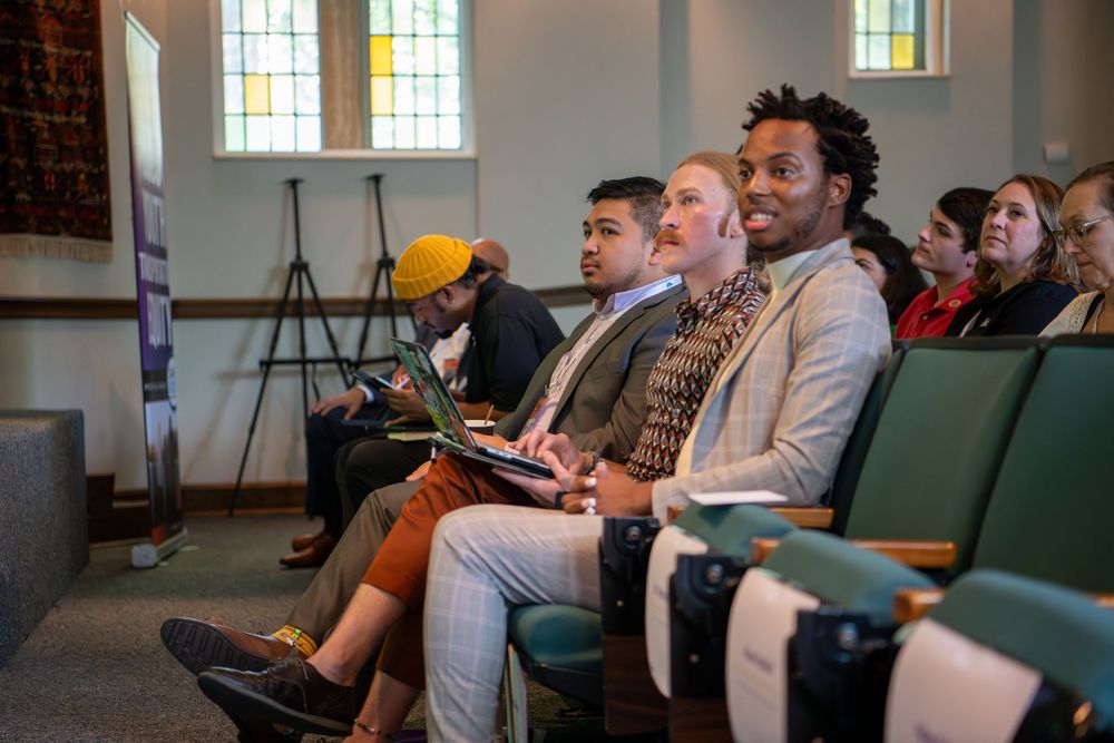 Diverse audience members sit attentively in a church or chapel setting with stained glass windows visible in the background.