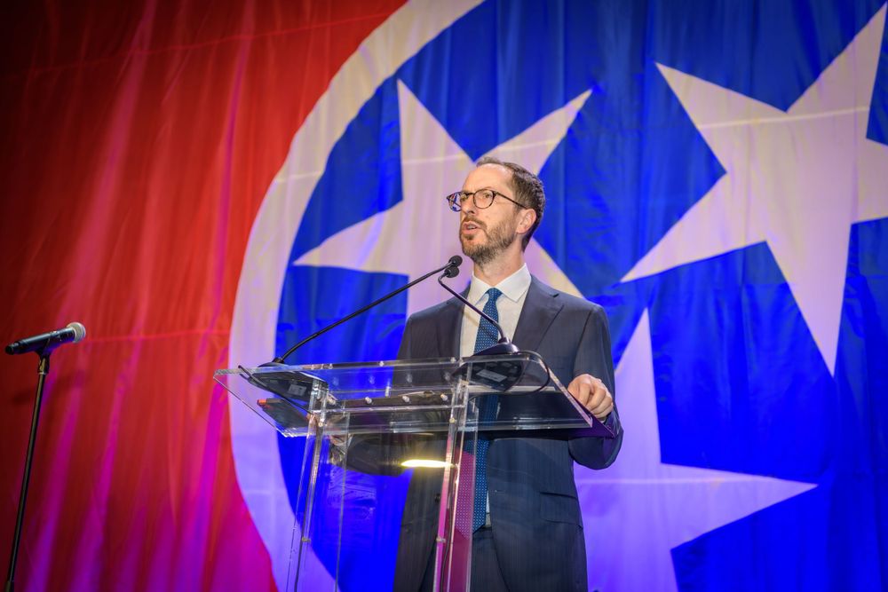A speaker stands at a transparent podium with a Tennessee state flag backdrop featuring red and blue colors with stars.