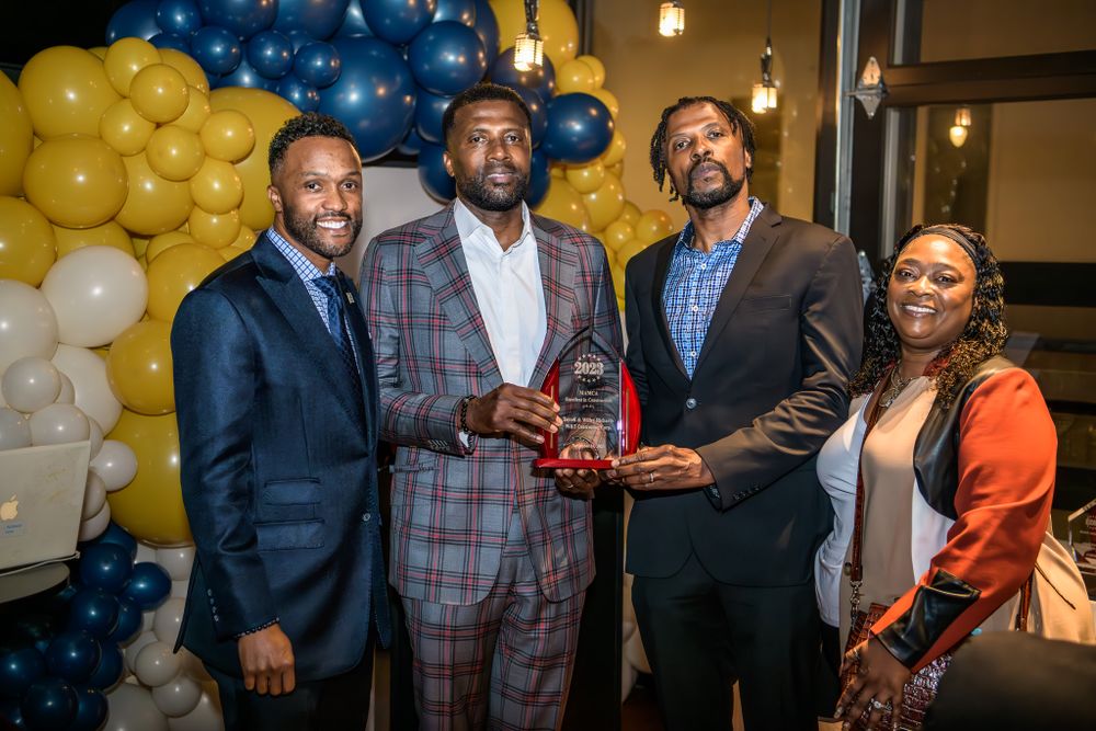 A group of people at a formal event pose together in front of yellow and blue balloon decorations.