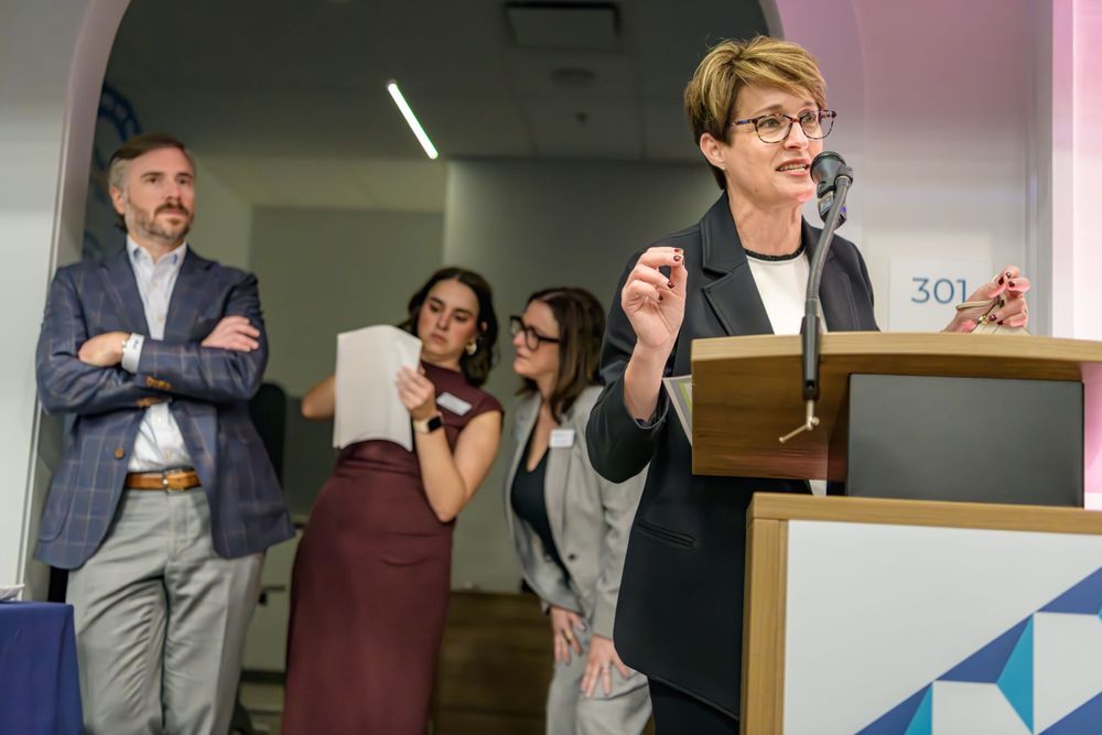 A speaker gestures at a podium while three onlookers stand nearby in an office setting.