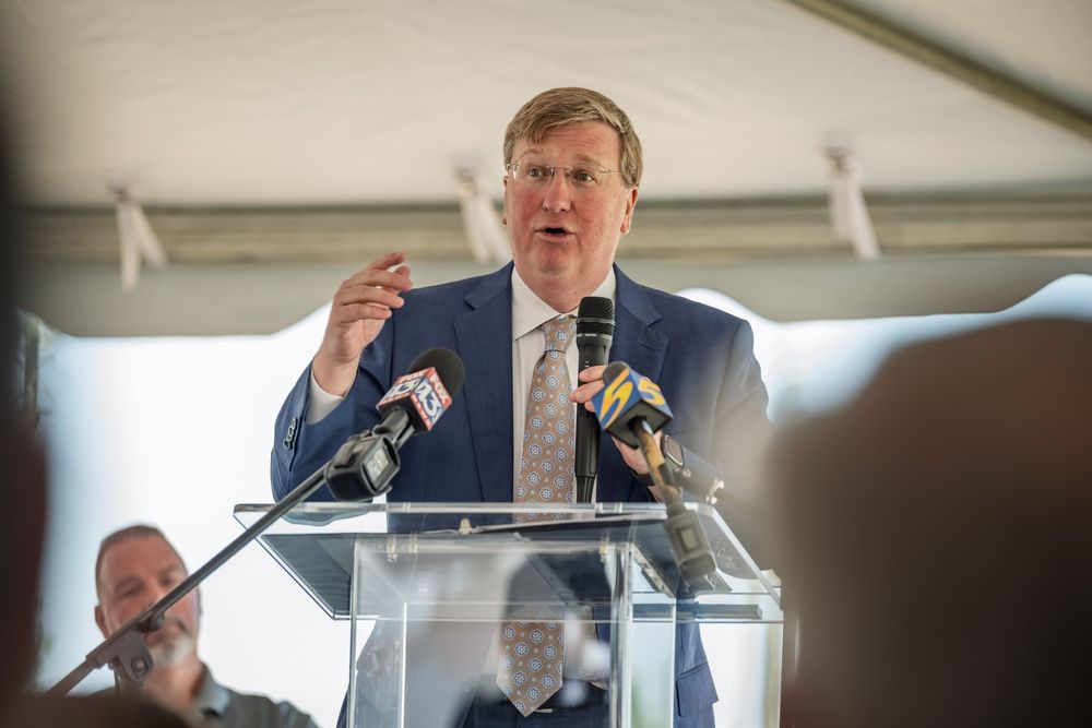 A man in a blue suit speaks passionately at a podium with microphones during an outdoor event under a tent.
