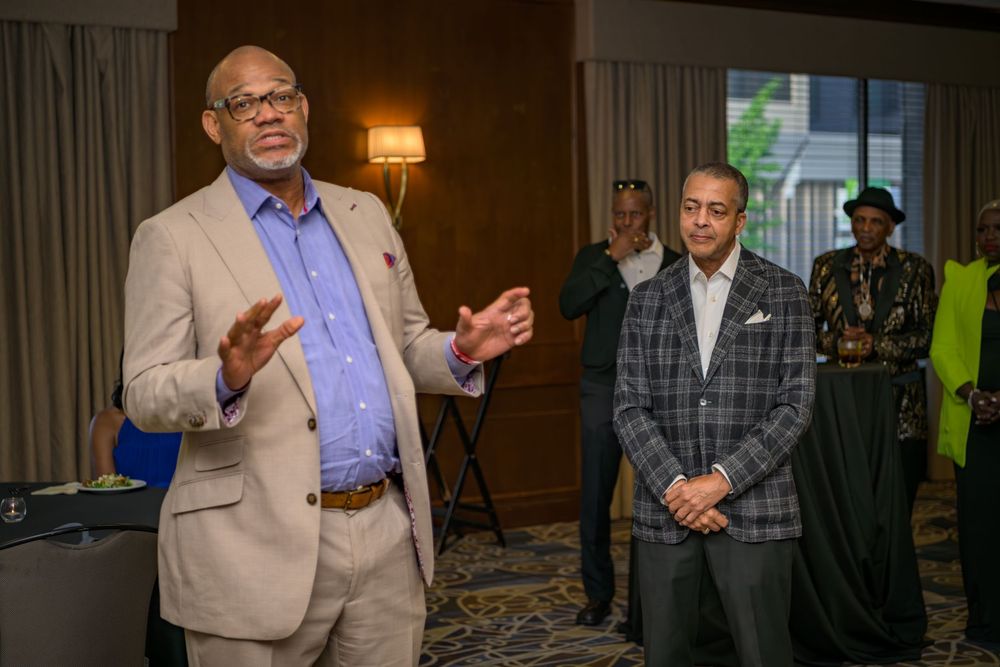 A professional speaker wearing a beige suit gestures while presenting at a conference event.