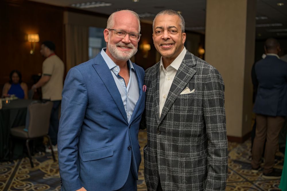 Two well-dressed men in blazers smile together at an indoor event with elegant lighting and decor.