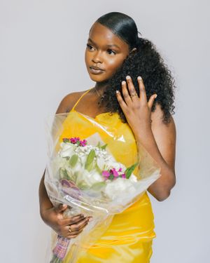 Young woman in yellow dress holding bouquet of flowers with curly side ponytail against white background.