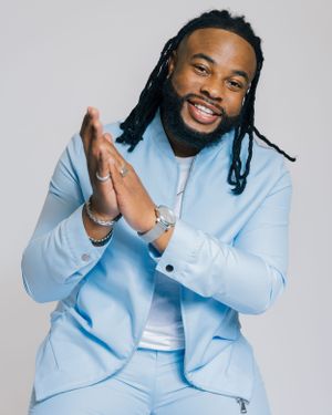 Man in light blue suit claps hands and smiles against grey background.