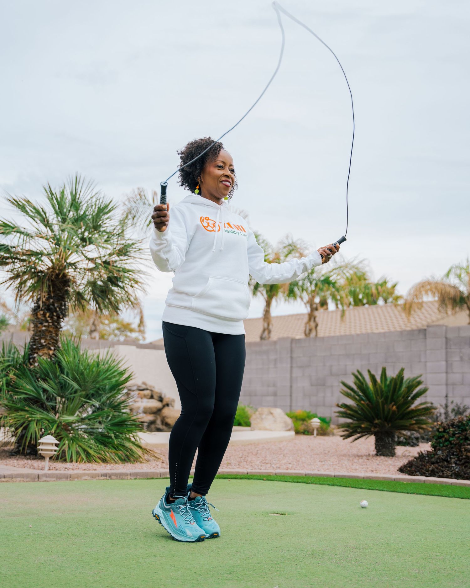 Woman in white sweatshirt jumps rope outdoors near palm trees.