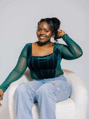 Woman in green mesh top and jeans poses casually on white chair.