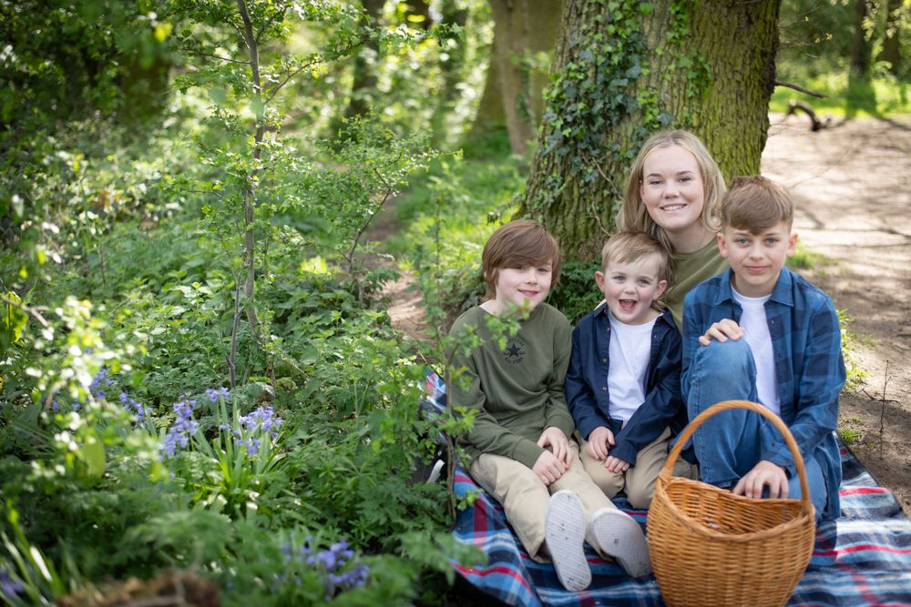 Children sitting on picnic blanket with wicker basket in woodland setting.