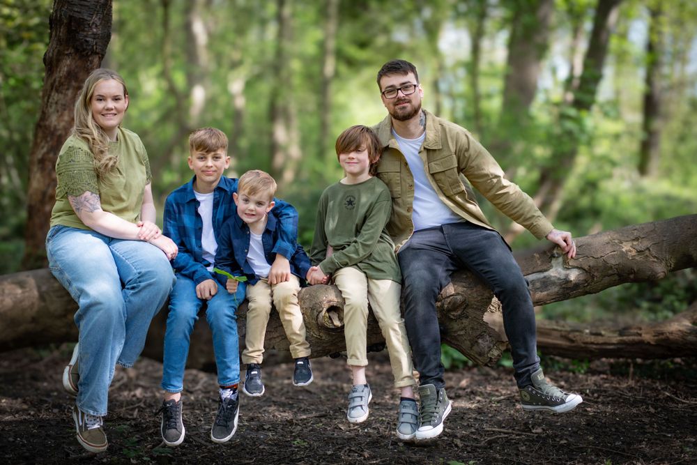 Family of five poses casually while sitting on large fallen log in woodland setting.