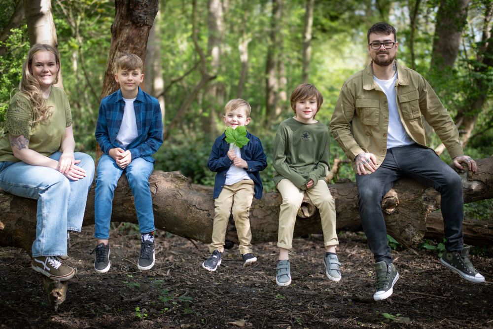Family group sits together on fallen tree trunk in wooded forest setting.