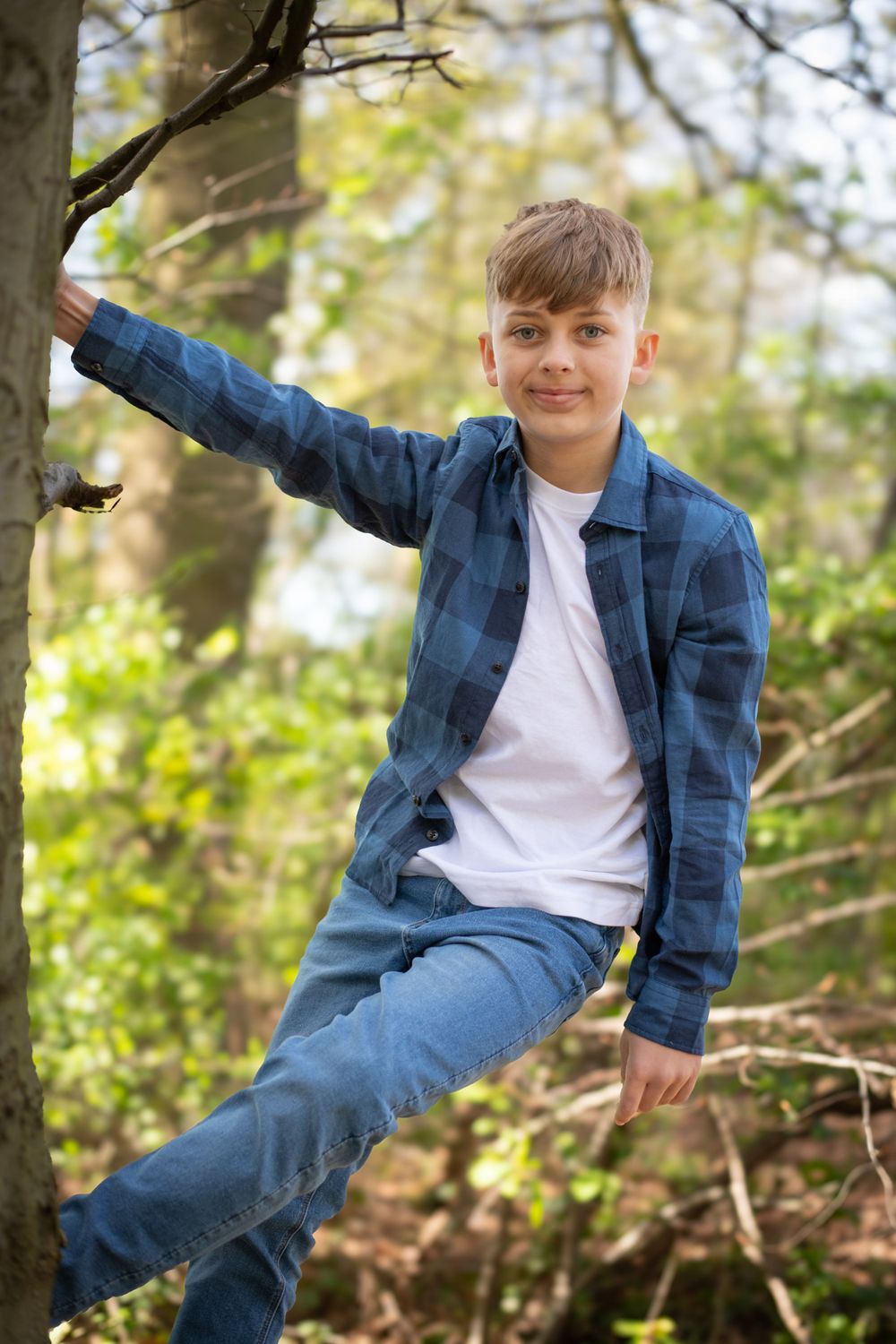 Youth in denim jacket rests casually on tree branch in sunny forest.