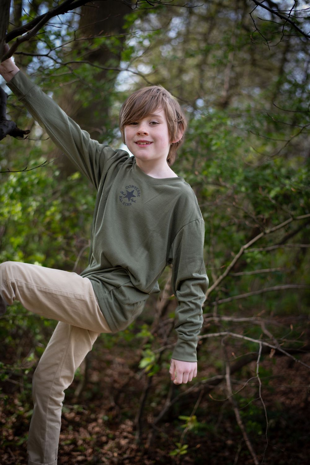 Young person in olive green sweatshirt playfully poses while climbing a tree in a forest.