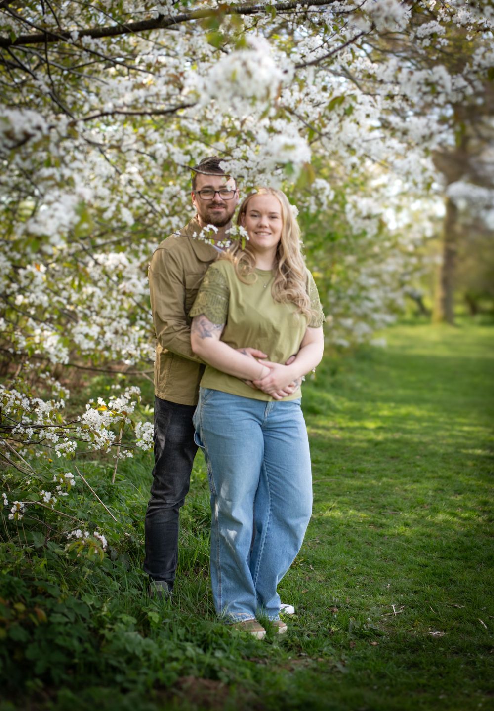 A couple poses under blooming spring cherry blossoms in a pastoral garden setting.