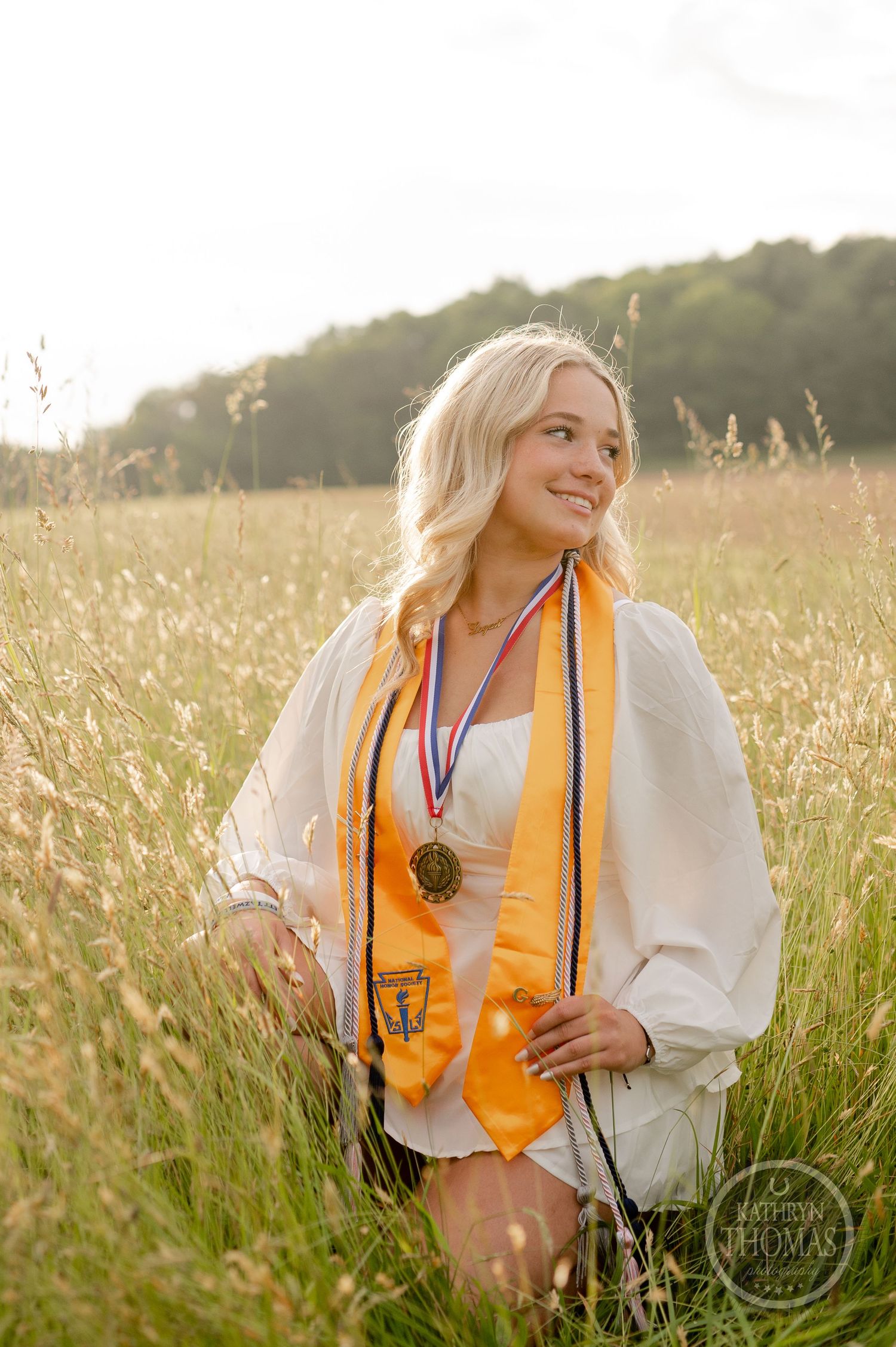 Graduate in white dress and gold stole poses in sunlit summer meadow.