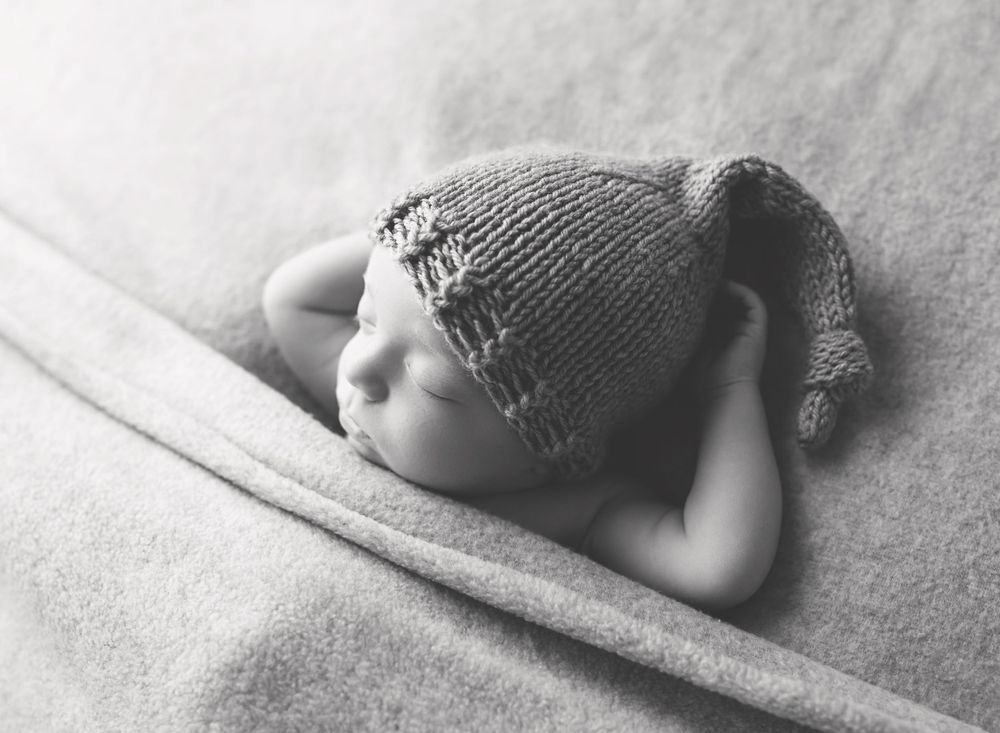 black and white photo of a newborn baby tucked in with arms above head wearing a knit cap