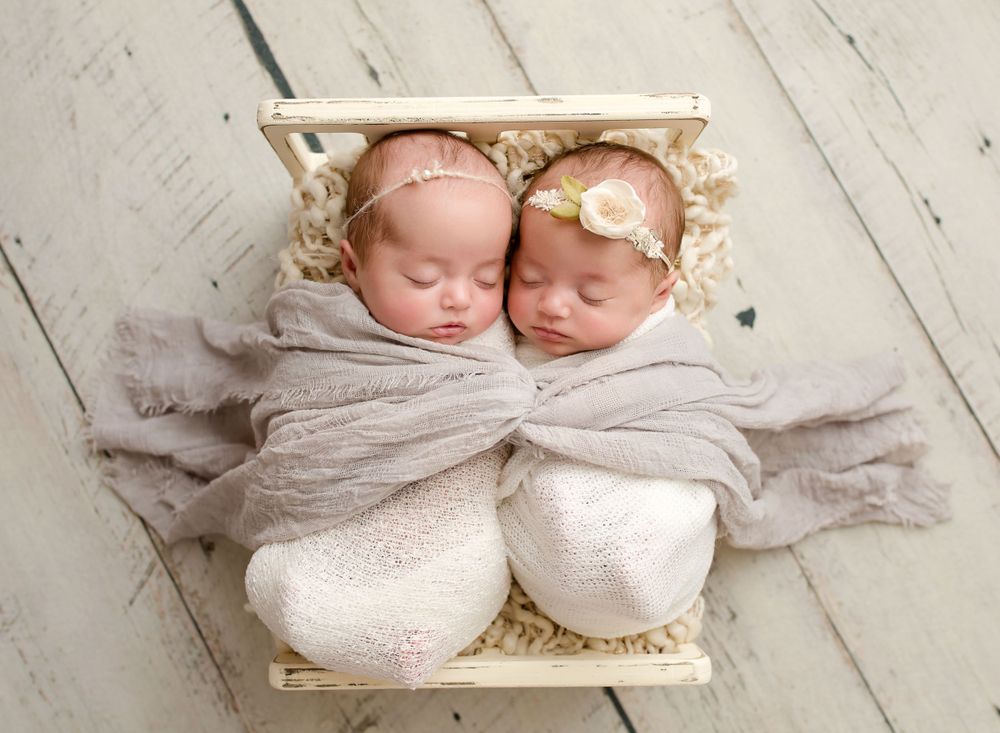 twin newborn girls wrapped in a white and gray wrap sleeping in a bed prop