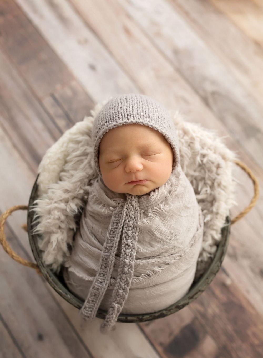 sleeping newborn baby wrapped like a burrito in a gray wrap with gray bonnet in a bucket on a wood backdrop