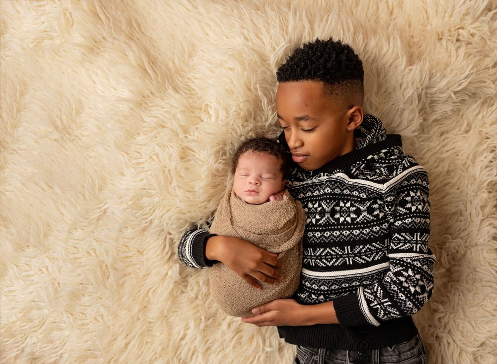 overhead photo of a big brother holding his newborn baby brother on a cream rug