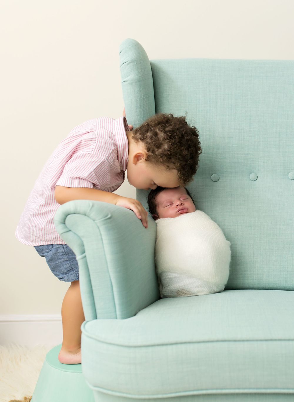 toddler boy leaning down to kiss newborn brother in a turquoise chair