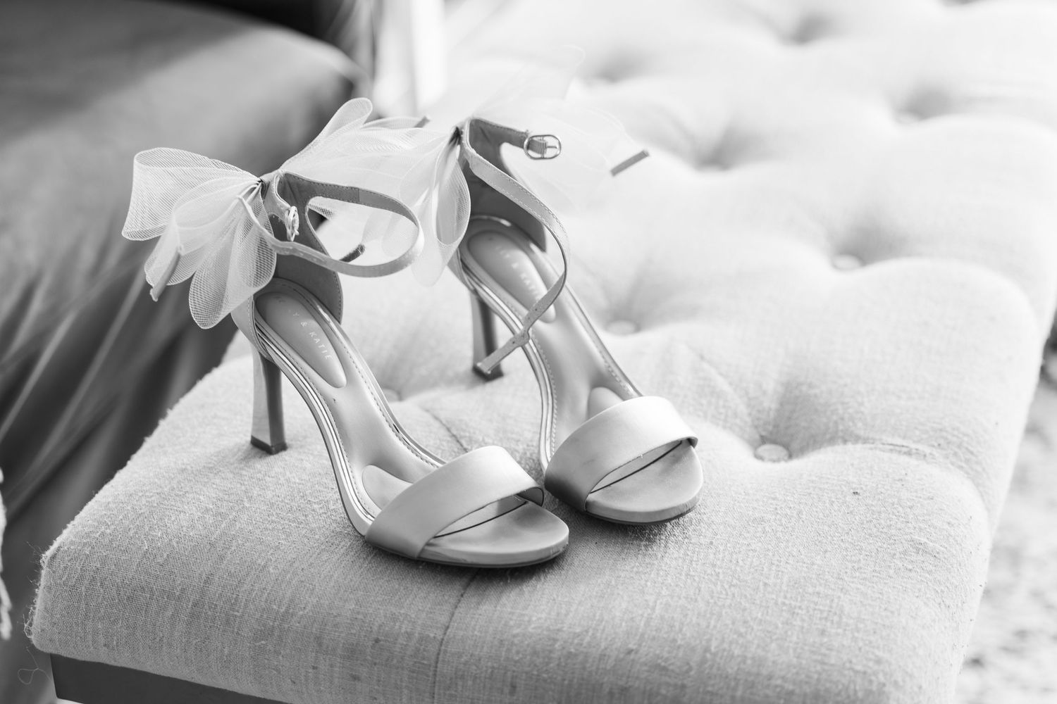 A pair of elegant white strappy high heel wedding shoes with bow detail resting on a tufted ottoman.