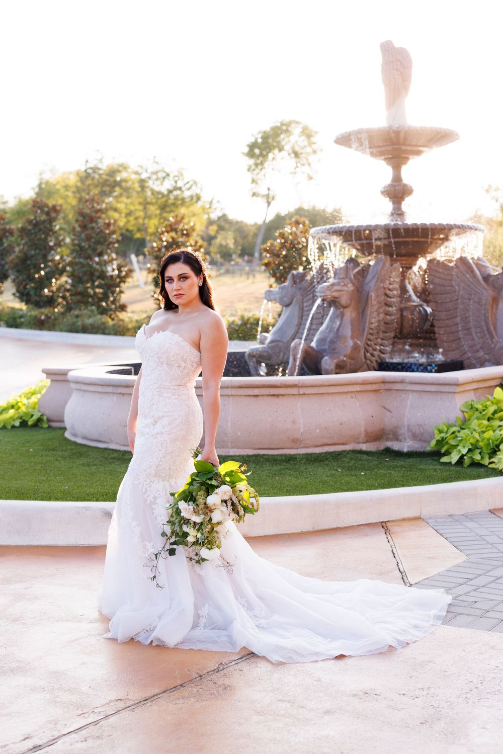 Elegant bridal session by ornate fountain featuring full white wedding gown and trailing bouquet.