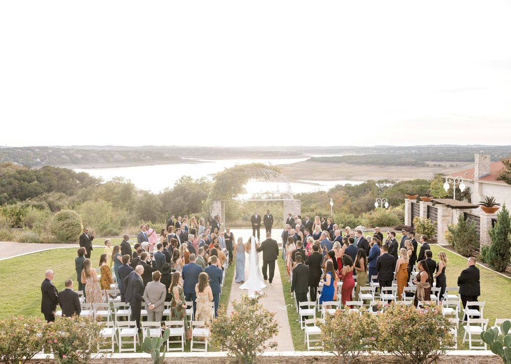 An outdoor wedding ceremony overlooking a scenic lake with guests seated in white chairs on a grassy hillside.